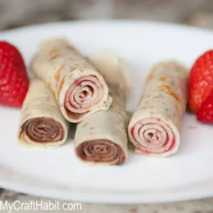 Crepes rolled with jam and nutella stacked on a white plate, surrounded by fresh strawberries