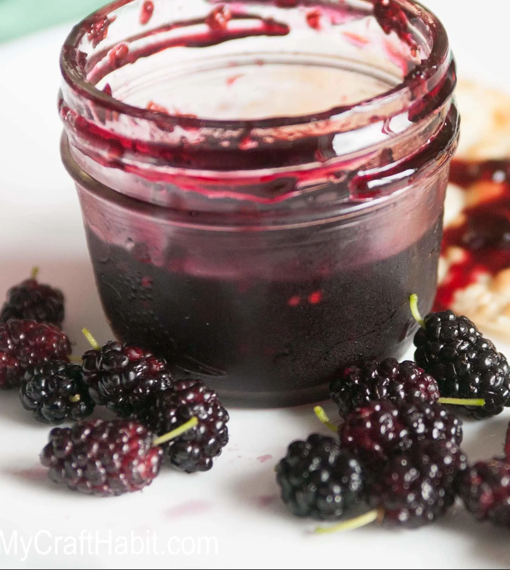 Small canning jar filled with homemade mulberry jam, surrounded by fresh mulberries on a white plate.