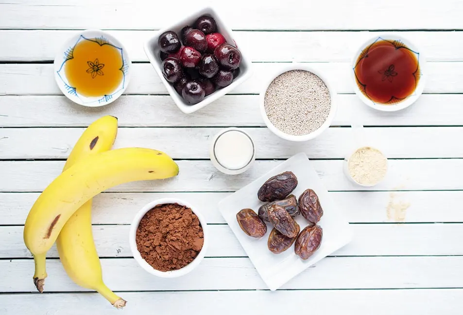 Ingredients to make a vegan chcoloate cherry bowl smoothie laid out on a white plank surface. Includes bananas, cherries, majool dates and seeds.