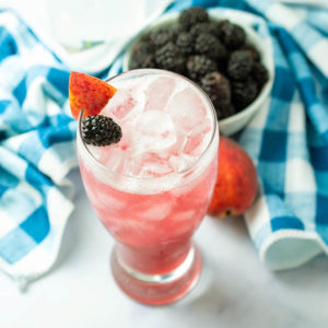 A tall glass filled with pink cocktail and garnished with a sliver of fresh peach and blackberry. A blue and white checkered napkin and small bowl of blackberries are in the background.