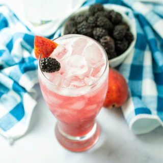 A tall glass filled with pink cocktail and garnished with a sliver of fresh peach and blackberry. A blue and white checkered napkin and small bowl of blackberries are in the background.
