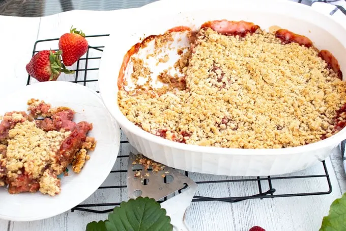 A white Corningware dish filled with baked strawberry and rhubarb crisp. On large piece has been taken out and put on a white plate. Fresh strawberries surround the dishes.