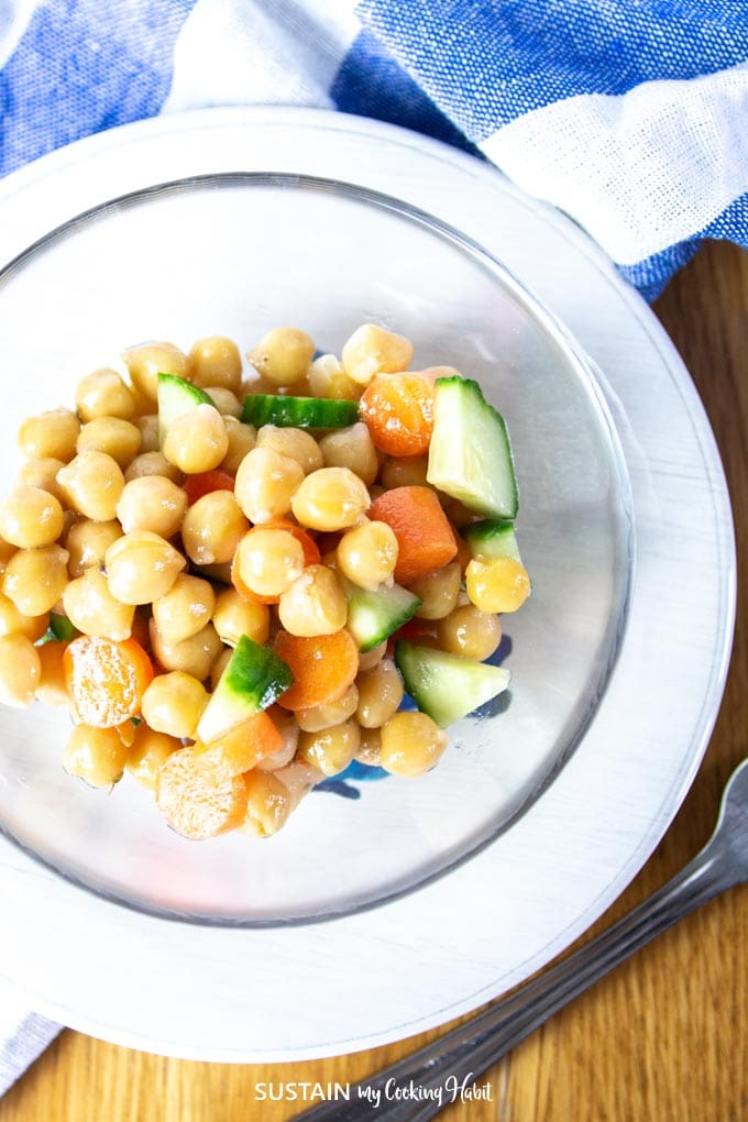 Chickpea salad with cucumbers and carrots in a clear bowl on a white plate. A blue checkered napkin is in the background.