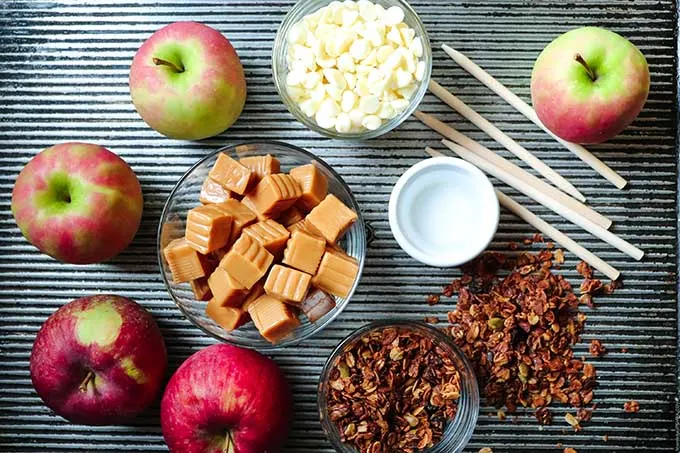 An overhead view of supplies needed to make the granola-dipped caramel apples recipe. Includes apples and small clear bowls filled caramels, white chocolate and granola on a dark grey surface. 