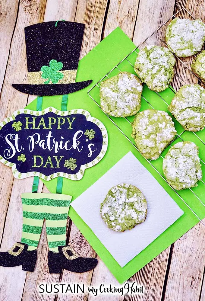 Finished and baked green crinkle cookies cooling on a cooling rack on a bright green place mat and rustic wood surface. A decorative sign reading Happy St. Patrick's Day is placed beside the cookies.