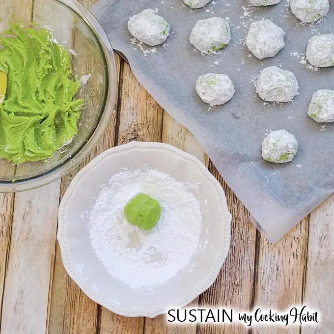A round green ball of dough being rolled in a place filled with powdered sugar. A cookie sheet filled with cookies ready to be baked is beside the plate.