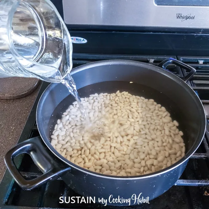 A pot on the stove filled with water-soak white beans. Additional water is being poured into the pot.