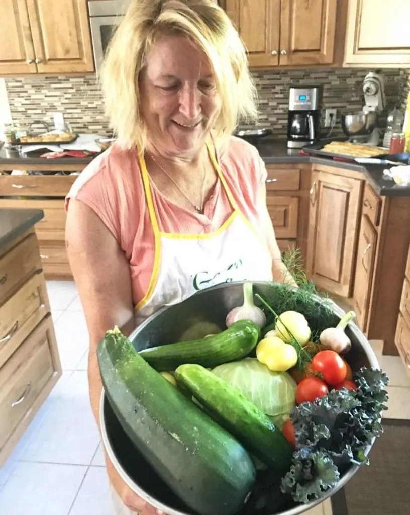 Woman holding a large silver bowl filled with cucumbers, tomatoes, kale, zuchinni and other vegetables.