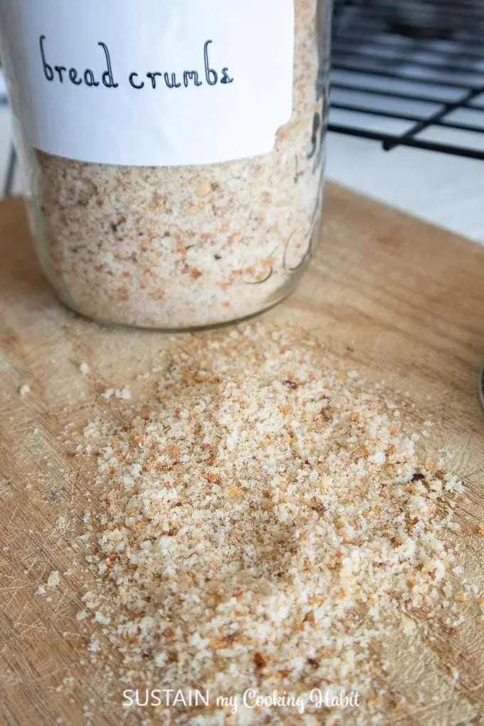 Close up image of homemade breadcrumbs on a wood cutting board surface. A labelled glass jar filled with bread crumbs is in the background.