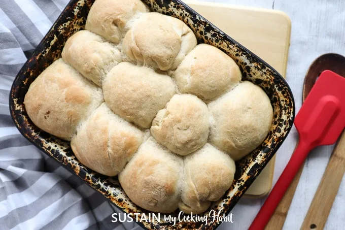 Overhead view of baked dinner rolls in a deep dish, placed next to a  cloth, spatula, cutting board and wooden spoon.