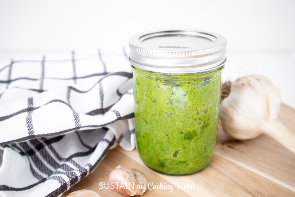 Garlic scapes pesto in a jar next to fresh garlic and a hand towel.