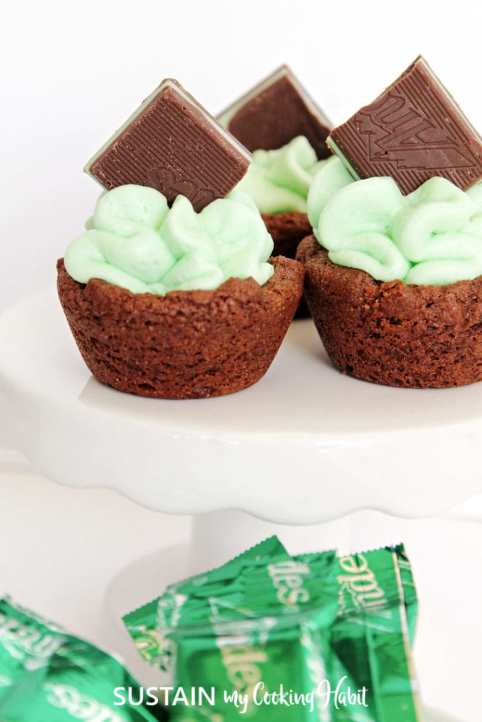 Chocolate mint cups with green frosting placed on a cake stand next to chocolate wrappers.