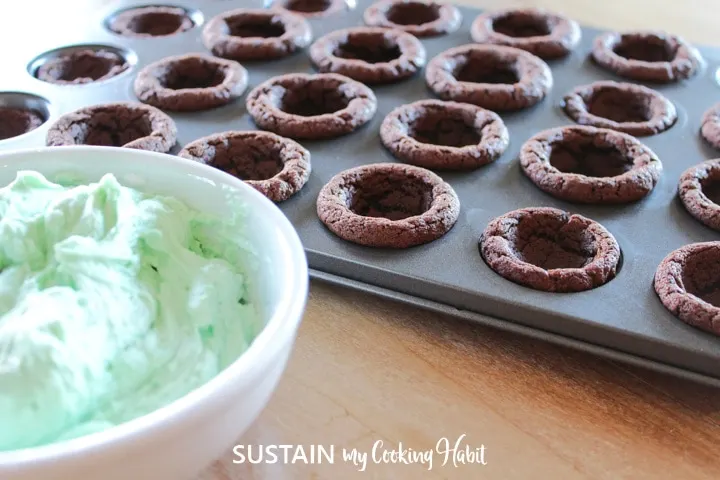 Green frosting in a bowl next to baked cookie cups.