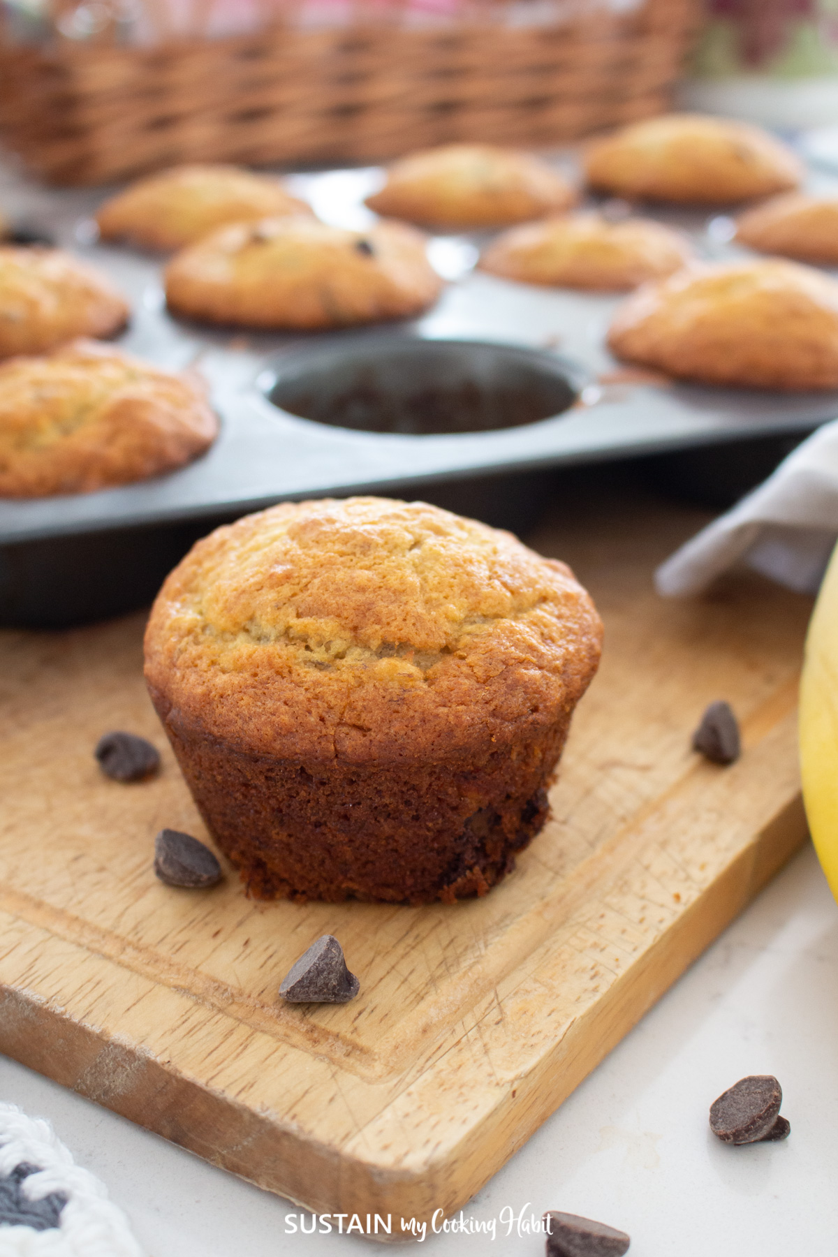 Banana chocolate chip muffin next to a pan of muffins and chocolate chips.