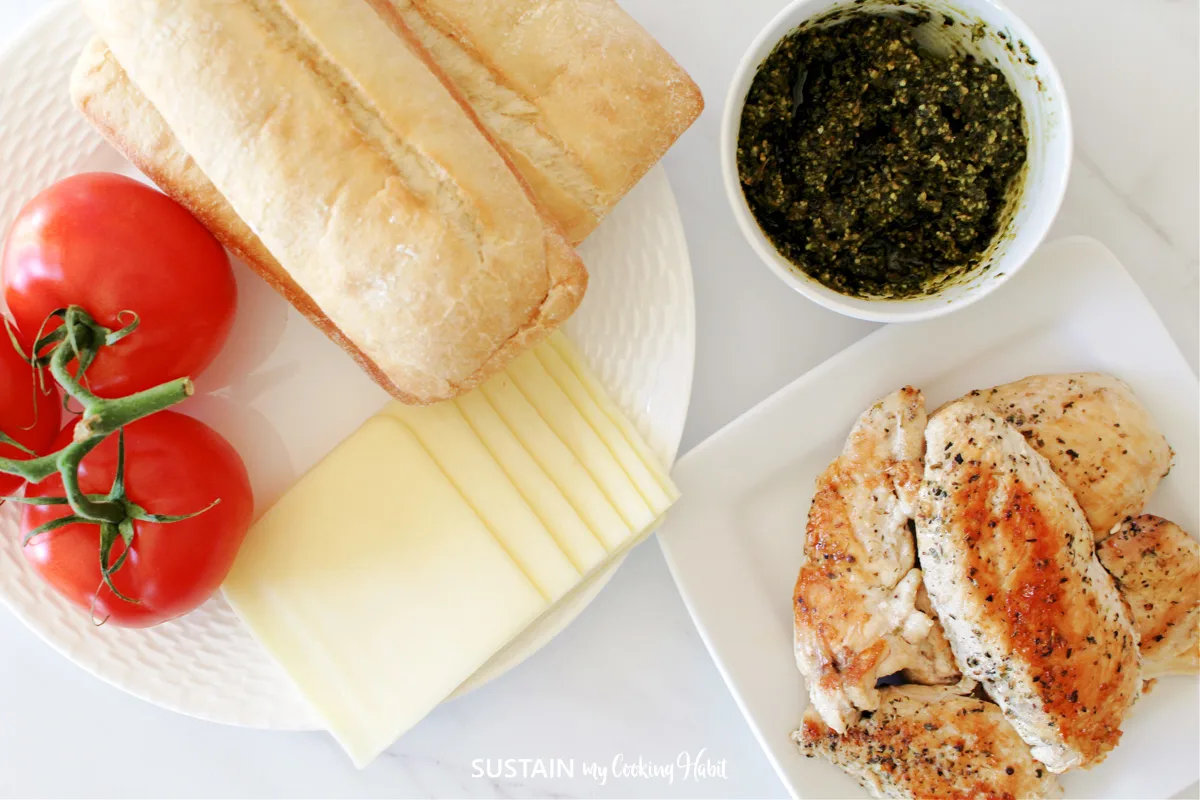 Ingredients need to make a chicken pesto panini including buns, chicken, pesto, cheese slices and tomatoes.